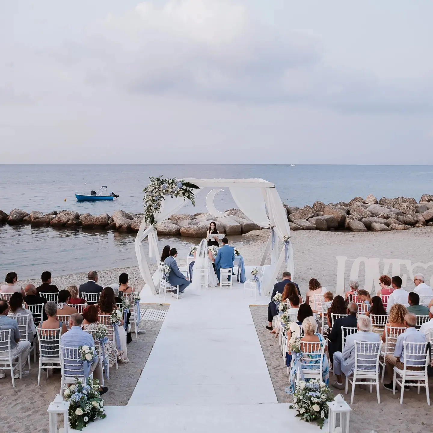 Cerimonia in spiaggia in Italia con gli ospiti seduti lungo una navata bianca Cerimonia in spiaggia in Italia con gli ospiti seduti lungo una navata bianca
