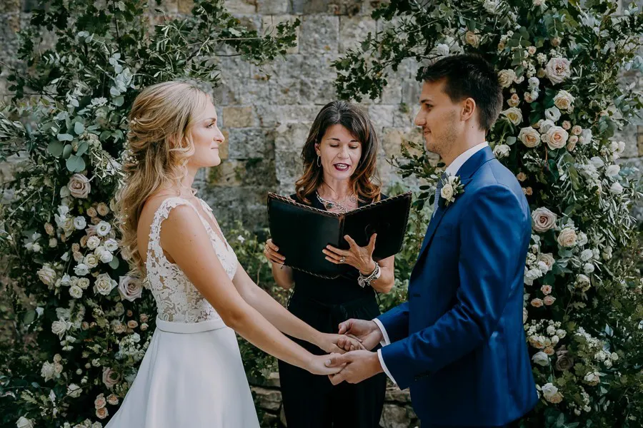 Couple holding hands with their celebrant under a floral installation by castle walls Couple holding hands with their celebrant under a floral installation by castle walls