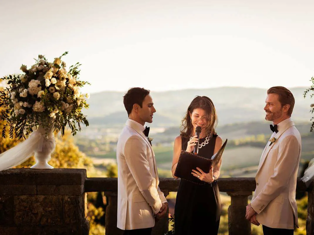 Photo: D2 Photography Two grooms in white dinner jackets at their wedding ceremony on a terrace overlooking the Tuscan hills