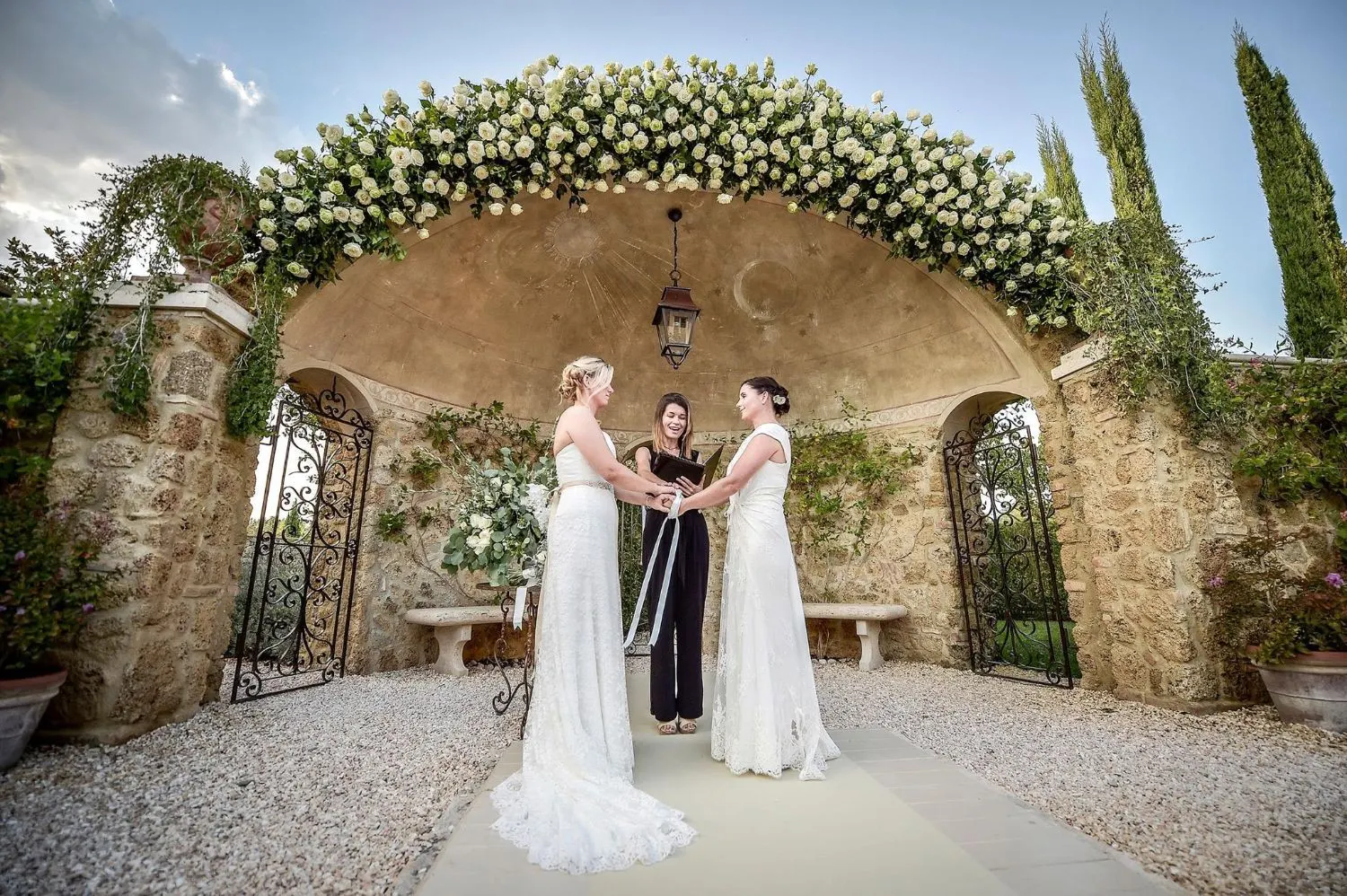 LGBTQ+ weddings in Italy Two brides during a handfasting ritual with their celebrant under a white rose arch