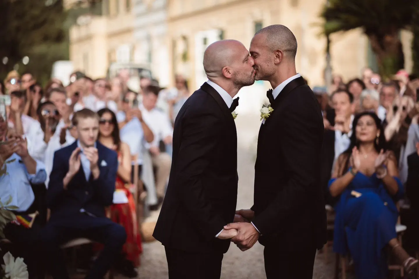 Photo: Giacomo Morelli Two grooms kissing at their wedding ceremony as guests applaud